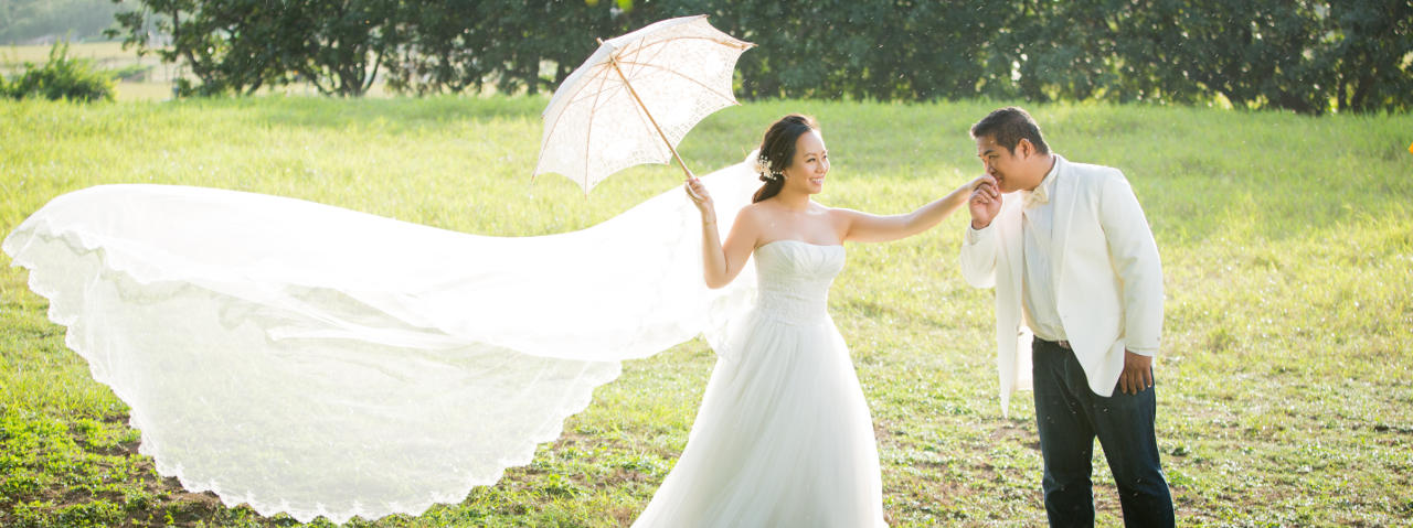 Nadia in a White Trailing Wedding Dress Holding and Umbrella in One Hand and the Other Being Kissed by JM in a White Tuxedo Jacket and Dark Blue Jeans Under an Umbrella in the Rain by Ching Hua Bridal Art